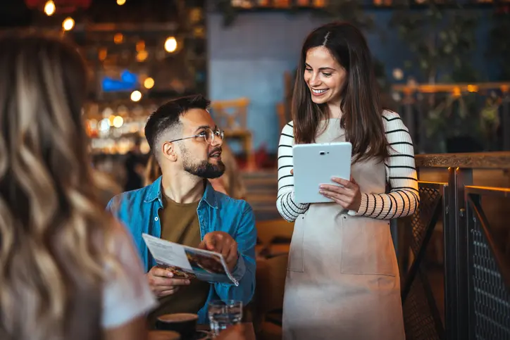 Goś w restauracji trzyma menu i składa zamówienie kelnerce, która trzyma tablet w ręku.